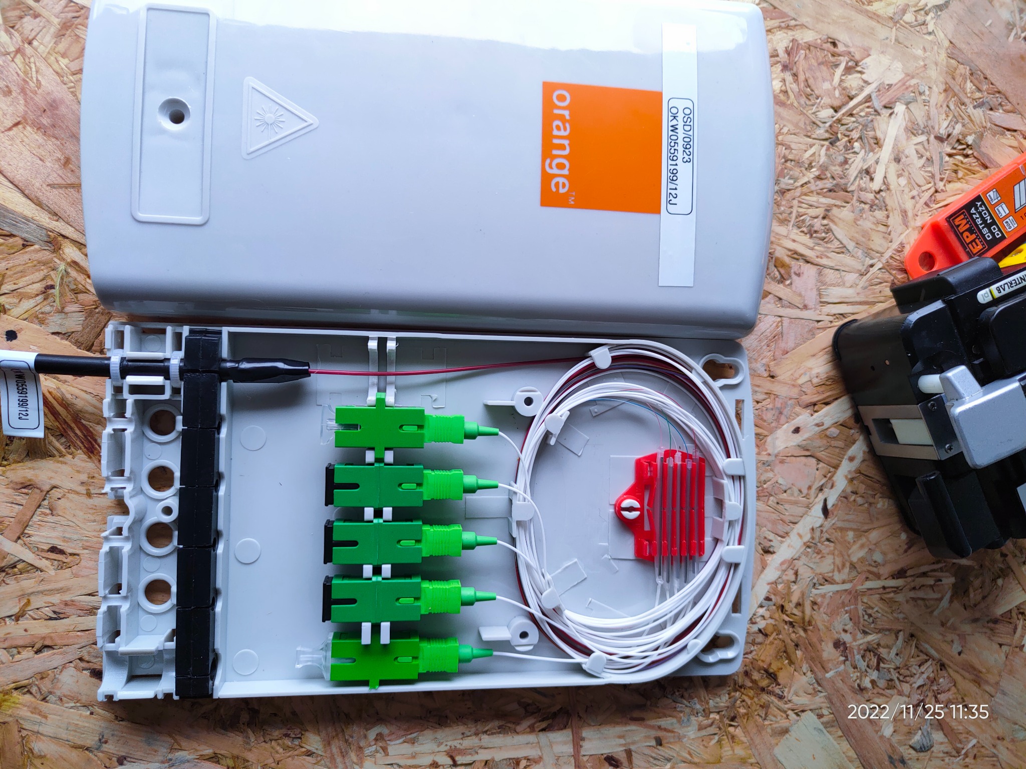 Otwarta skrzynka telekomunikacyjna Orange z podłączonymi światłowodami i narzędziem do spawania światłowodów na drewnianym podłożu.