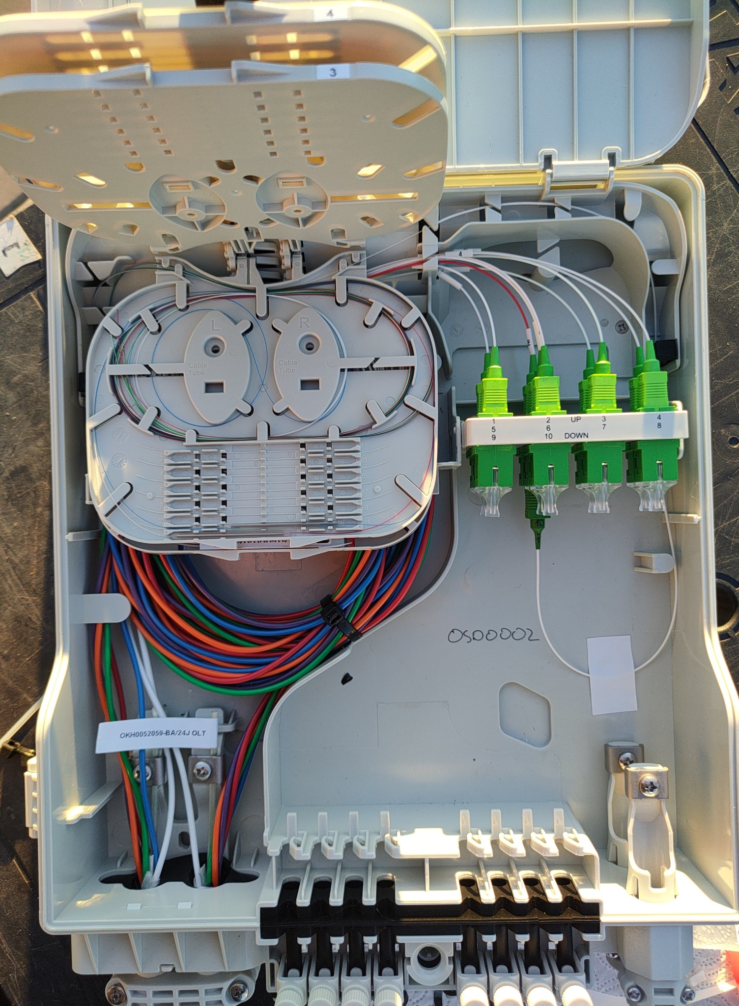 Otwarta skrzynka telekomunikacyjna z kolorowymi kablami światłowodowymi i zielonymi złączkami, widok z góry, etykieta z kodem OKH0052059-BA/24J OLT.