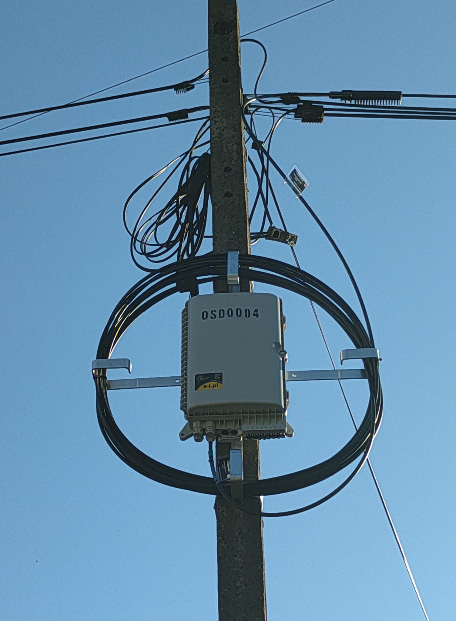 Słup telekomunikacyjny z zamontowaną skrzynką OSD0004 i okablowaniem. Widok z dołu na tle błękitnego nieba.
