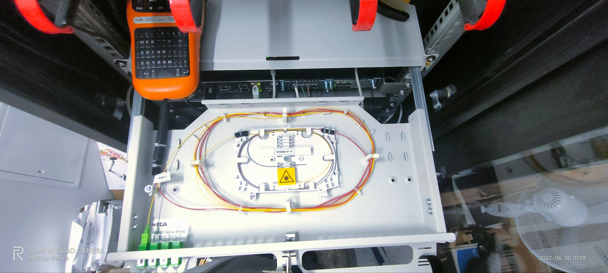 Panel światłowodowy z okablowaniem, widoczne złącza i kable światłowodowe w kolorach żółtym i pomarańczowym. Na górze tester kabli z klawiaturą.
