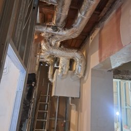 Klimatyzacja w Krakowie Maciej Kowalczyk - Montaż rekuperacji: srebrne rury wentylacyjne pod sufitem, biała centrala na ścianie, drabina i pracownik w tle. Widoczne elementy konstrukcyjne budynku.