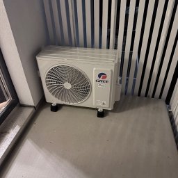 Klimatyzacja w Krakowie Maciej Kowalczyk - Biała jednostka zewnętrzna klimatyzacji marki Gree, umieszczona na małych kółkach, stoi na balkonie z białymi barierkami. Widoczne logo i tabliczka znamionowa.