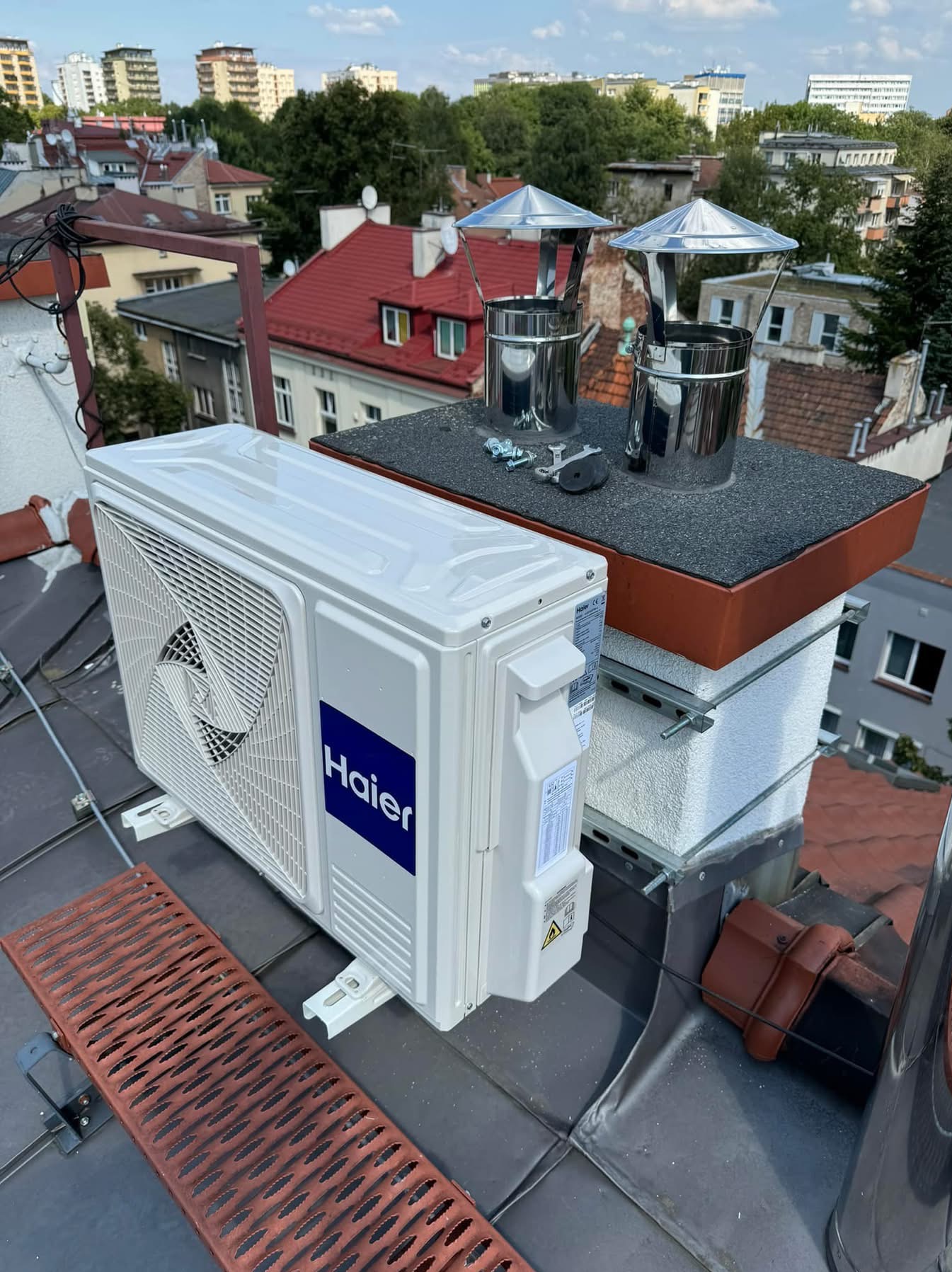 Biała jednostka zewnętrzna klimatyzacji Haier zamontowana na dachu budynku, obok komina z metalowymi nasadami. W tle zabudowa miejska i błękitne niebo.