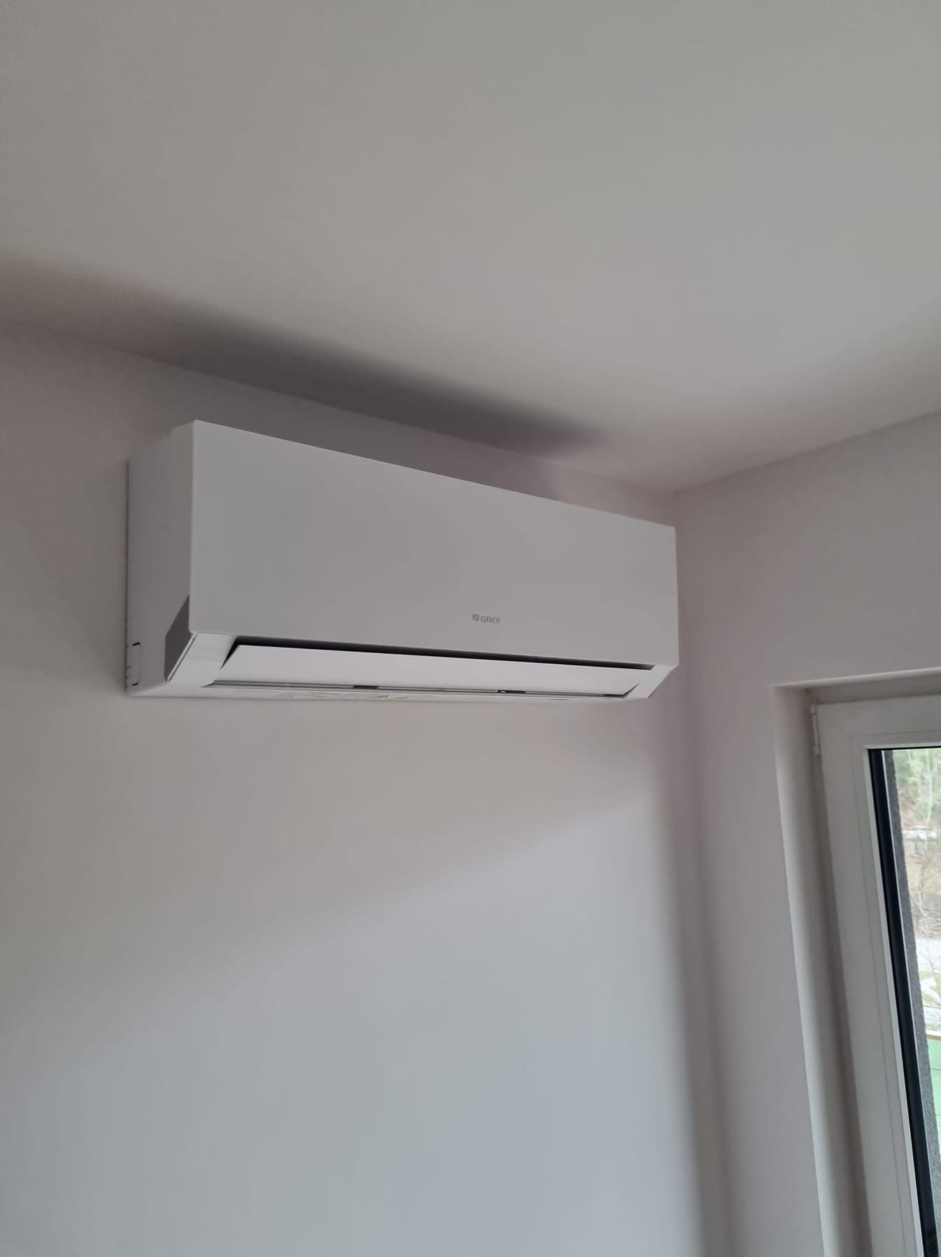 Biały klimatyzator marki Gree zamontowany na jasnej ścianie w pomieszczeniu, widoczny fragment okna po prawej stronie. Minimalistyczny design.