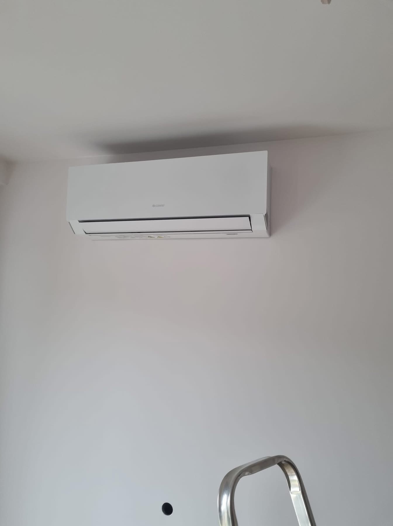 Biały klimatyzator ścienny marki Gree zamontowany w jasnym pomieszczeniu. Fragment drabiny w dolnej części kadru sugeruje niedawny montaż.