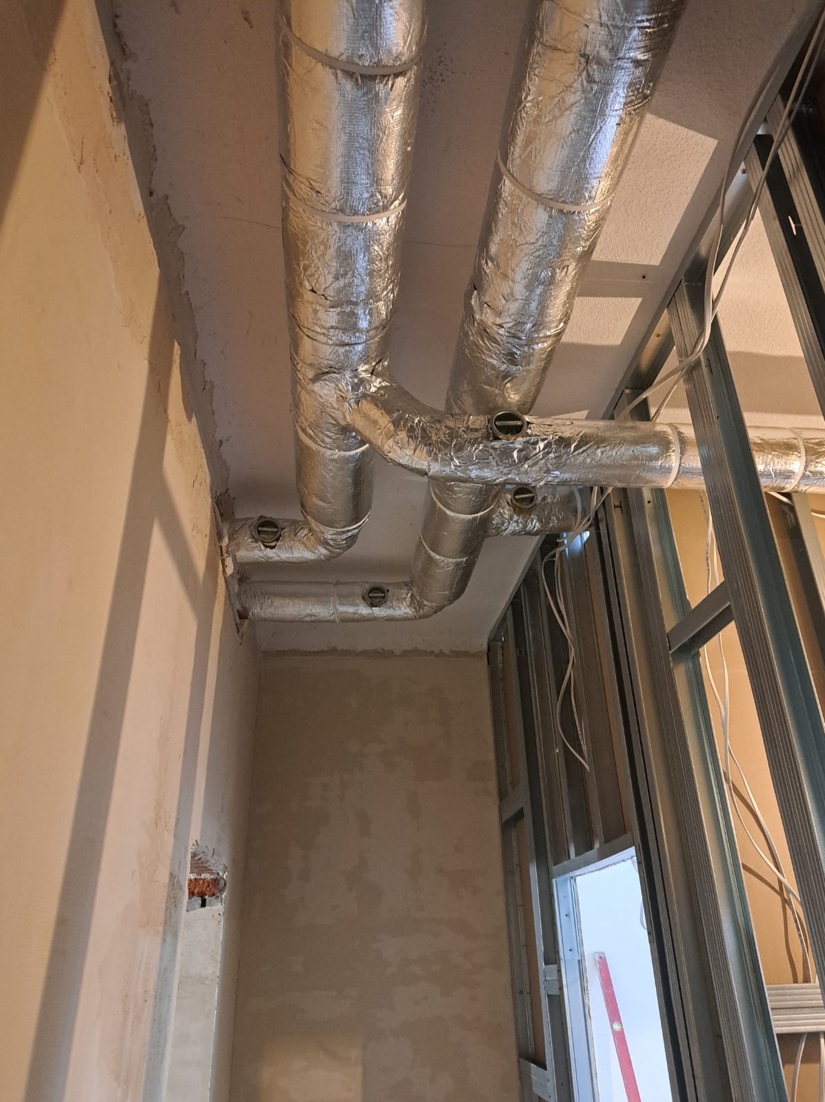 Montaż izolowanych przewodów wentylacyjnych pod sufitem, widoczne elementy konstrukcji metalowej i fragment ściany w surowym stanie.