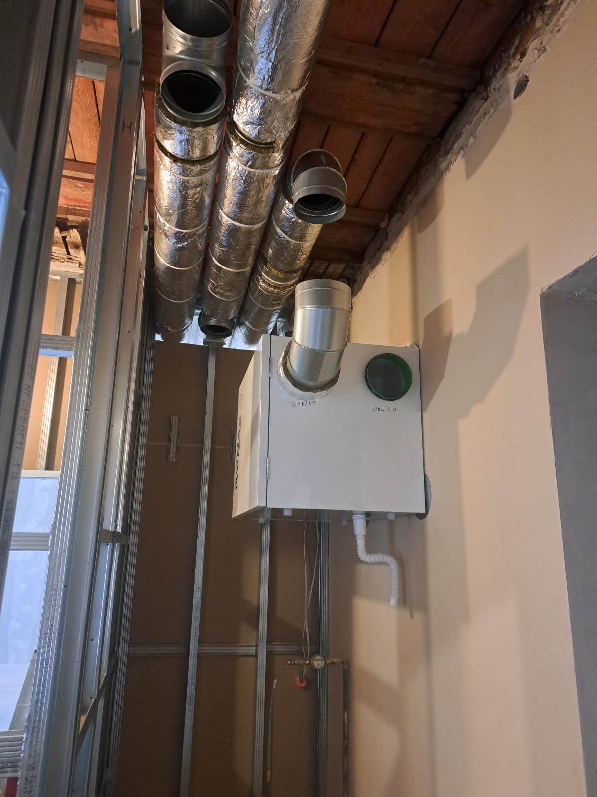 Montaż rekuperacji: biała jednostka centralna z rurami wentylacyjnymi w surowym wnętrzu, widoczne elementy konstrukcyjne ściany i drewniany strop.