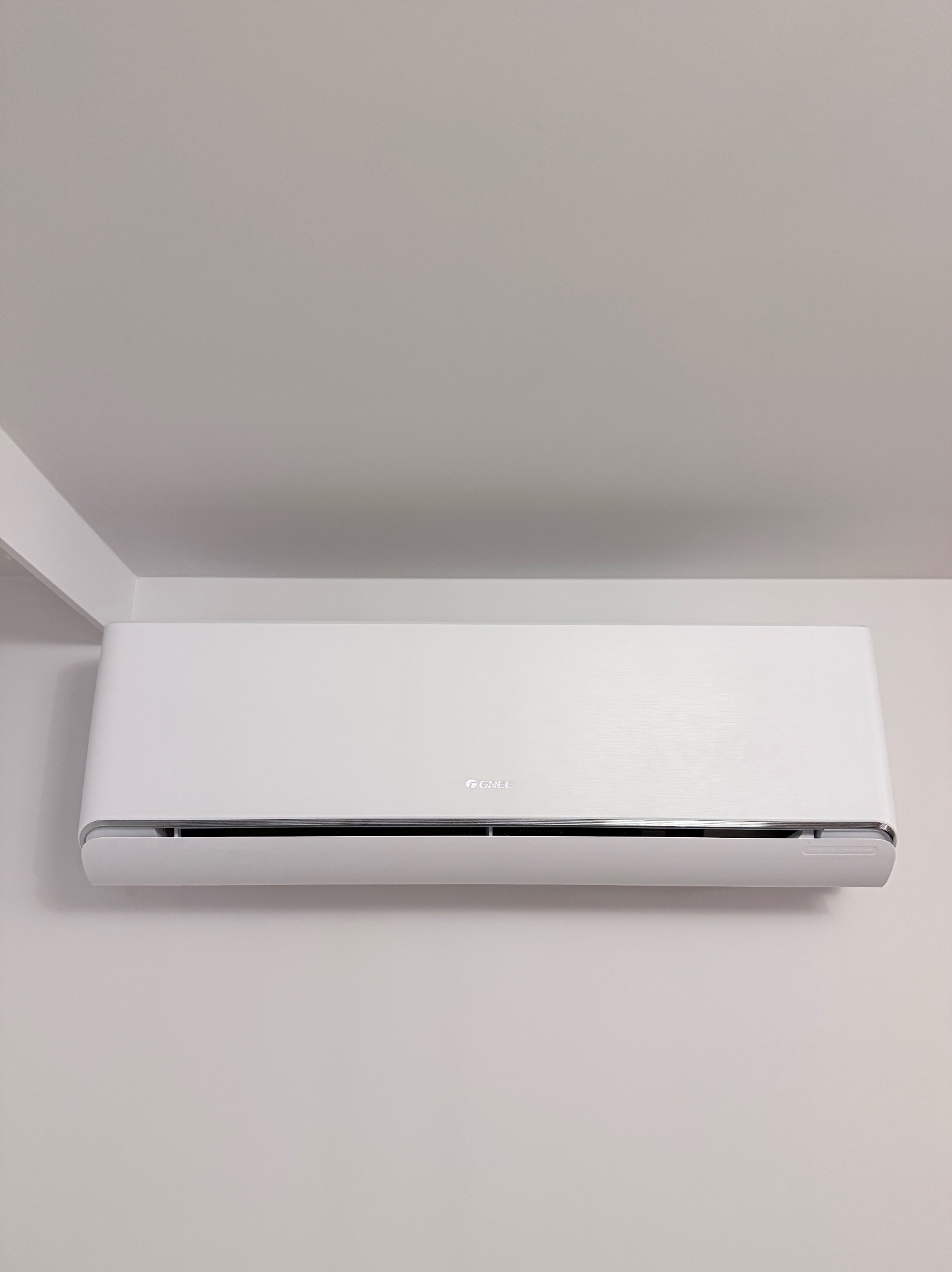 Biały klimatyzator marki Gree zamontowany na jasnej ścianie w pomieszczeniu. Widoczny fragment sufitu i narożnika ściany. Minimalistyczny design.