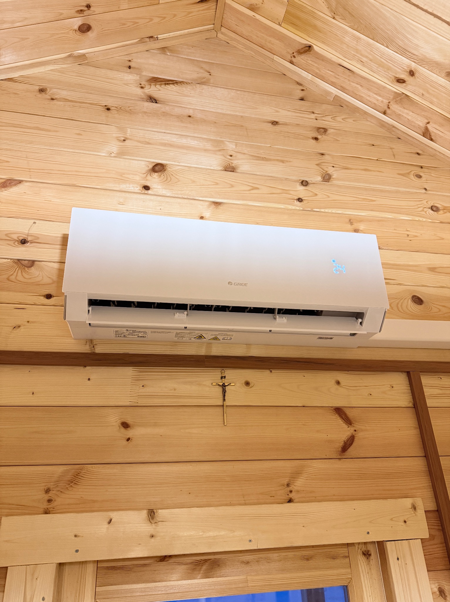 Biała klimatyzacja marki Gree zamontowana na drewnianej ścianie wewnątrz pomieszczenia, widok z dołu, krzyżyk wisi na ścianie.