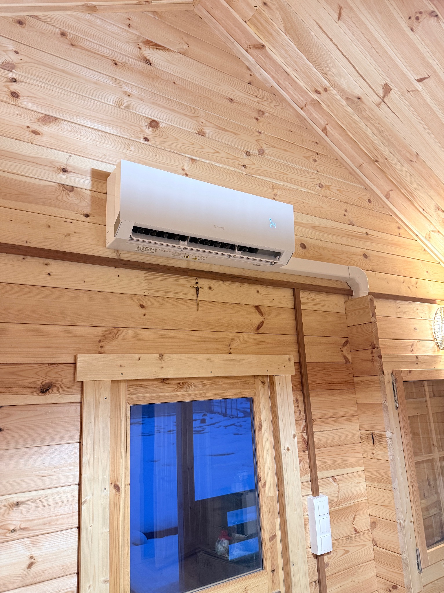 Biała klimatyzacja na drewnianej ścianie w domu z boazerią, widoczne okno i krzyżyk. Minimalistyczny design, jasne wnętrze.