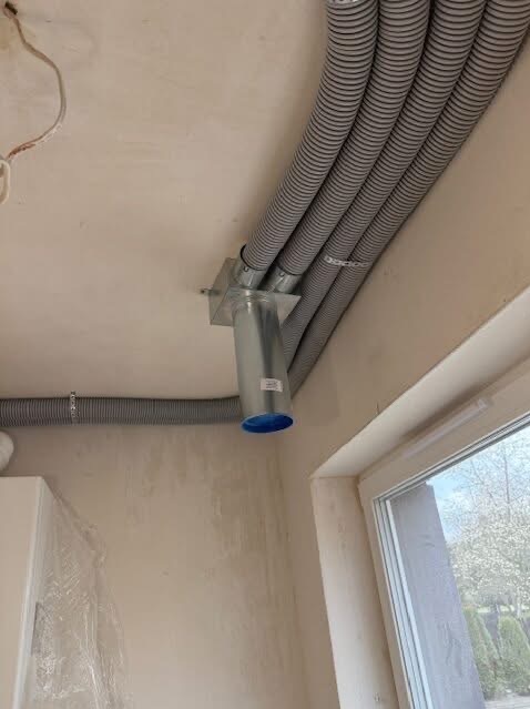 Szare, elastyczne rury wentylacyjne przymocowane do sufitu, z metalową puszką i niebieskim zakończeniem, widoczne okno w tle. Instalacja w trakcie montażu.