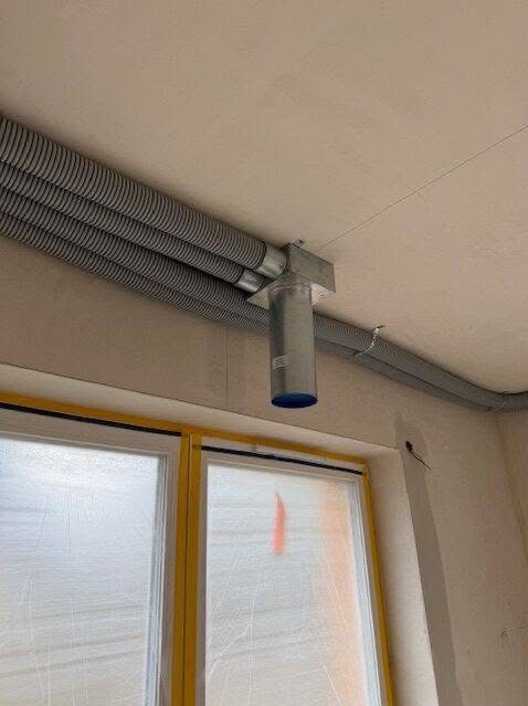 Szare, elastyczne rury wentylacyjne przymocowane do sufitu, z metalowym elementem łączącym. Okno zabezpieczone folią malarską w tle.