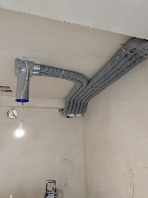 Szare, karbowane rury wentylacyjne zamontowane pod sufitem w surowym wnętrzu. Widoczny fragment instalacji z metalową puszką i wiszącą żarówką.