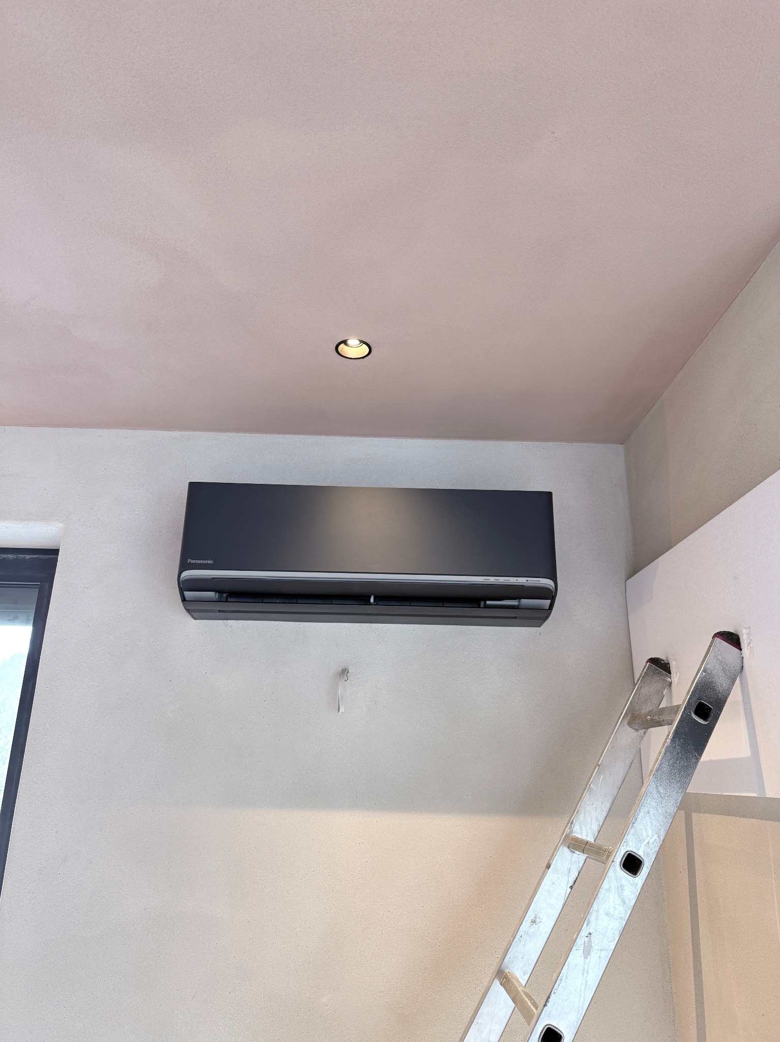 Czarny klimatyzator Panasonic zamontowany na ścianie w pomieszczeniu z drabiną, widok z dołu. Świeżo wykończone wnętrze, sufit w odcieniach różu.
