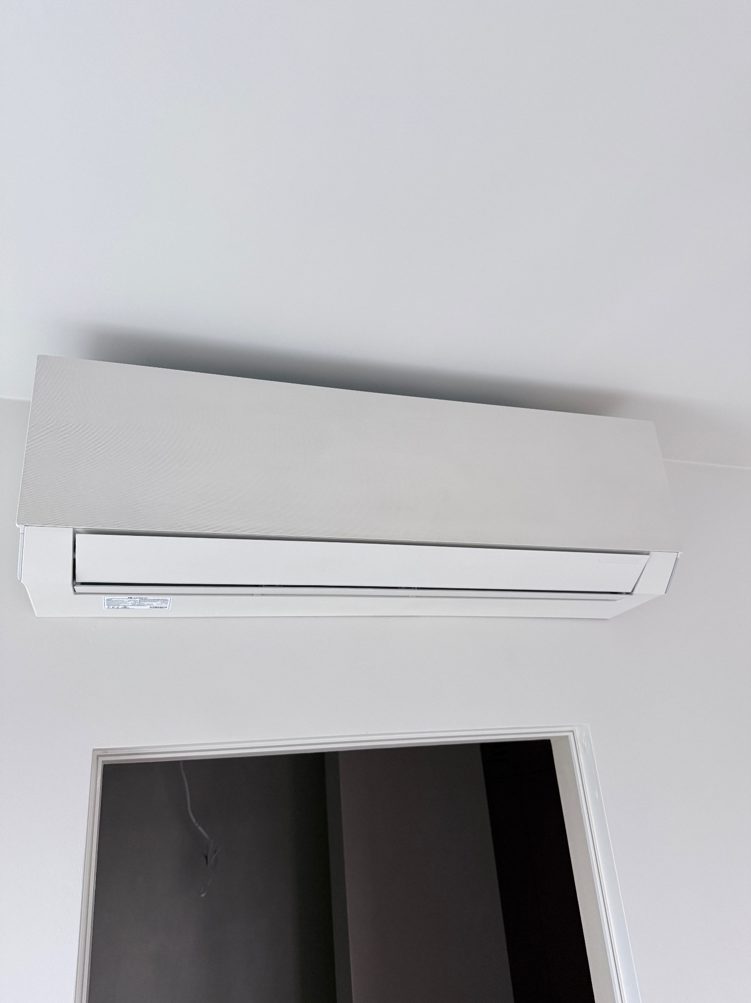 Biała klimatyzacja ścienna zamontowana nad drzwiami w minimalistycznym wnętrzu. Widoczny fragment sufitu i ściany w jasnych barwach.