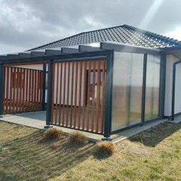 Radomir Wolny - Elegancka konstrukcja zewnętrzna z ciemnoszarego metalu, drewnianymi pionowymi lamelami i przezroczystymi panelami, tworząca zadaszone patio przy białym domu, na tle trawnika.