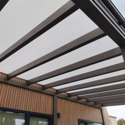 Radomir Wolny - Nowoczesna pergola z ciemnymi drewnianymi belkami i półprzezroczystym dachem z poliwęglanu, zamontowana nad drewnianą elewacją budynku z czarnymi oknami.