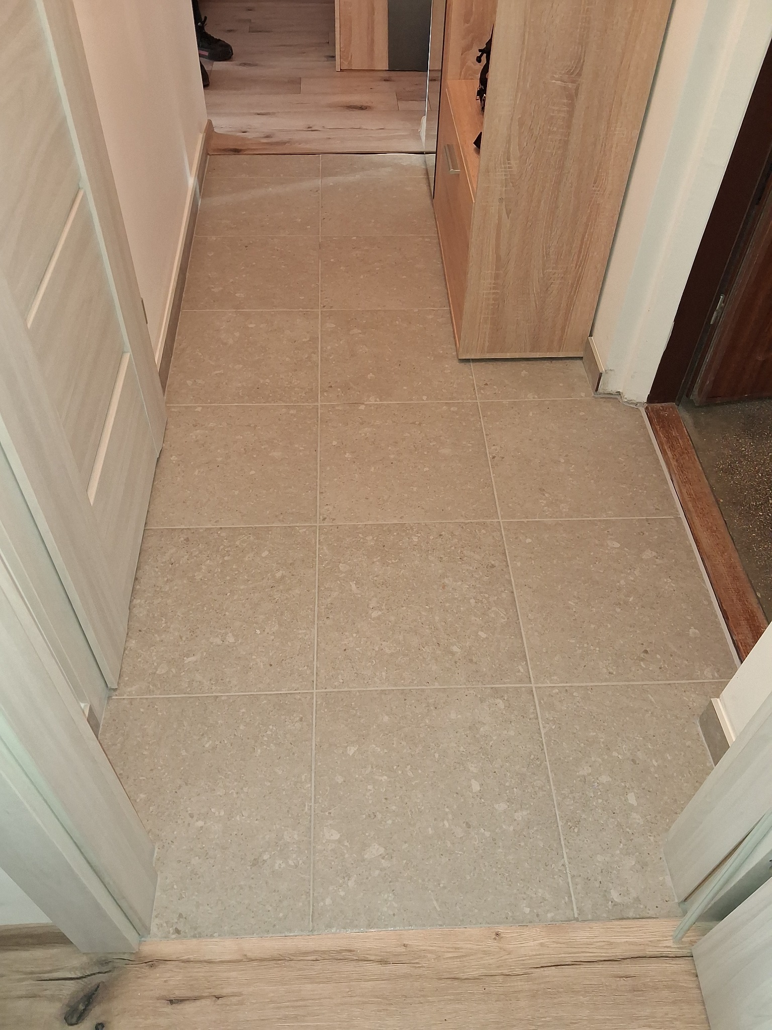 Beżowe, kwadratowe płytki podłogowe z delikatnym wzorem w korytarzu, widoczne drzwi i fragment szafki. Jasne fugi podkreślają regularny układ płytek.