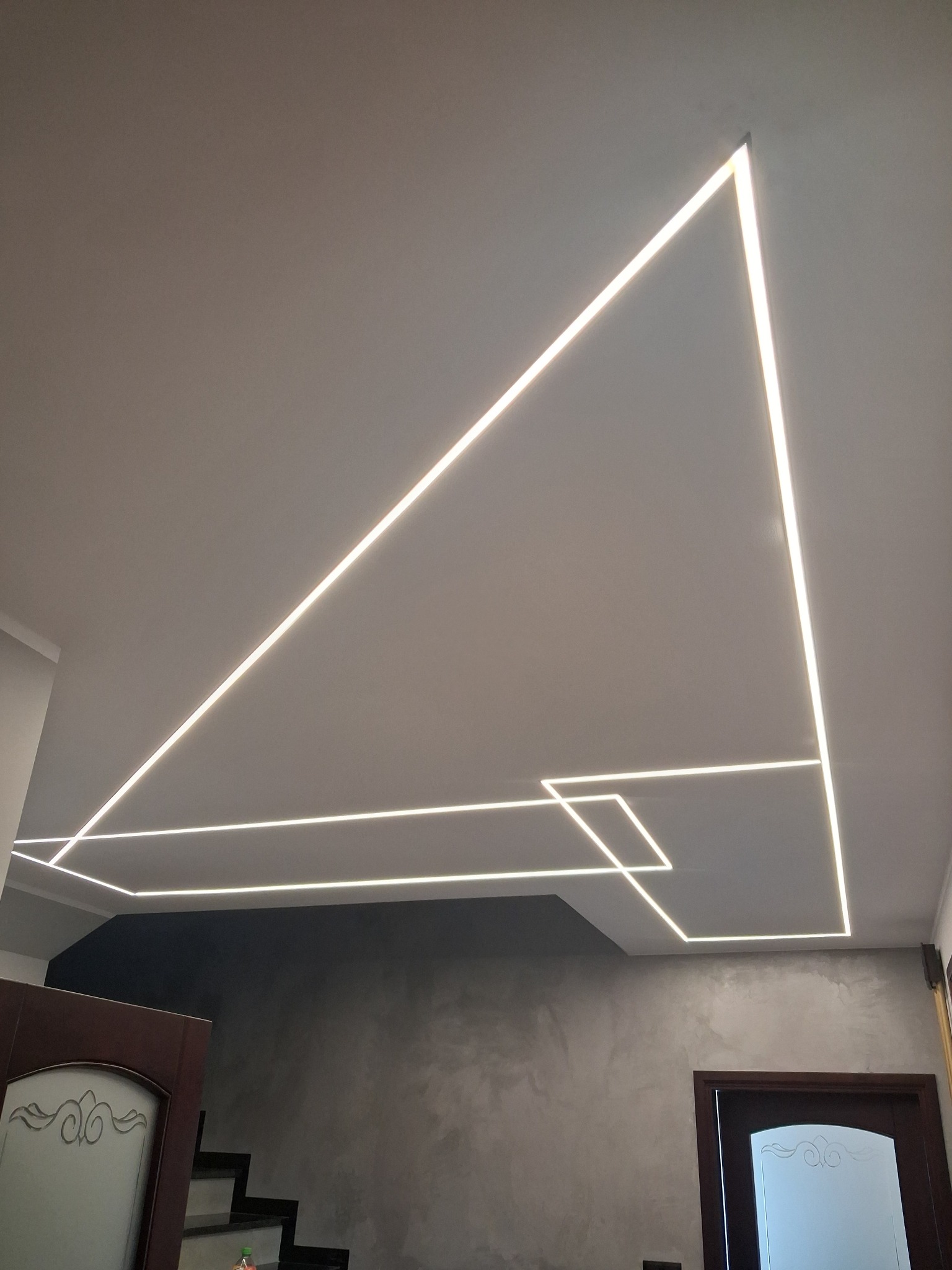 Nowoczesne oświetlenie sufitowe LED w kształcie geometrycznym, z widocznymi drzwiami i fragmentem schodów w tle. Minimalistyczny design wnętrza.