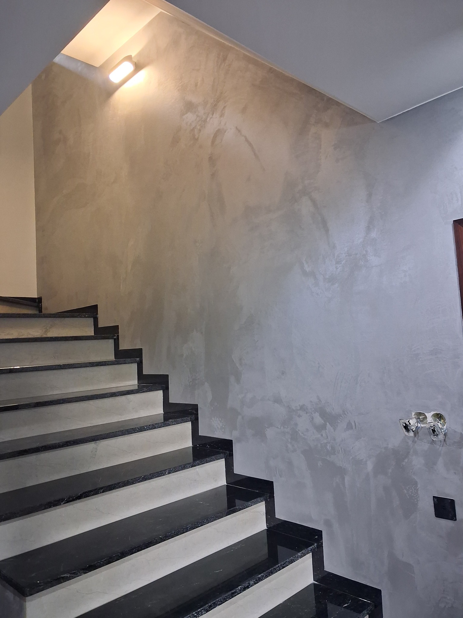 Schody z czarnym granitem i szara ściana z efektem dekoracyjnym w Skarszewach. Widoczne gniazdka elektryczne i oświetlenie schodowe.