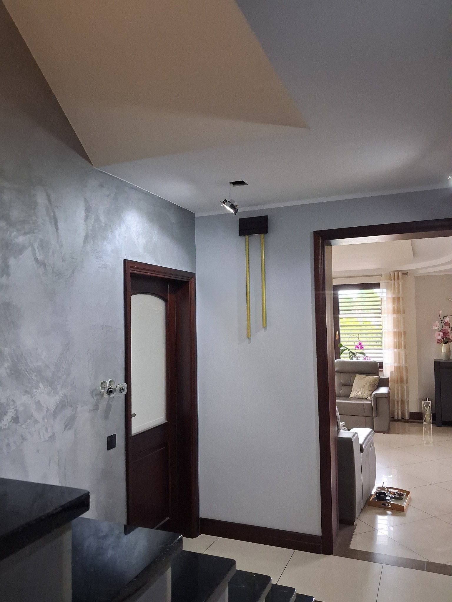 Stylowe wykończenie wnętrza: ściana z efektem dekoracyjnym w odcieniach szarości, drewniane drzwi i widok na salon z jasnymi zasłonami i sofą.