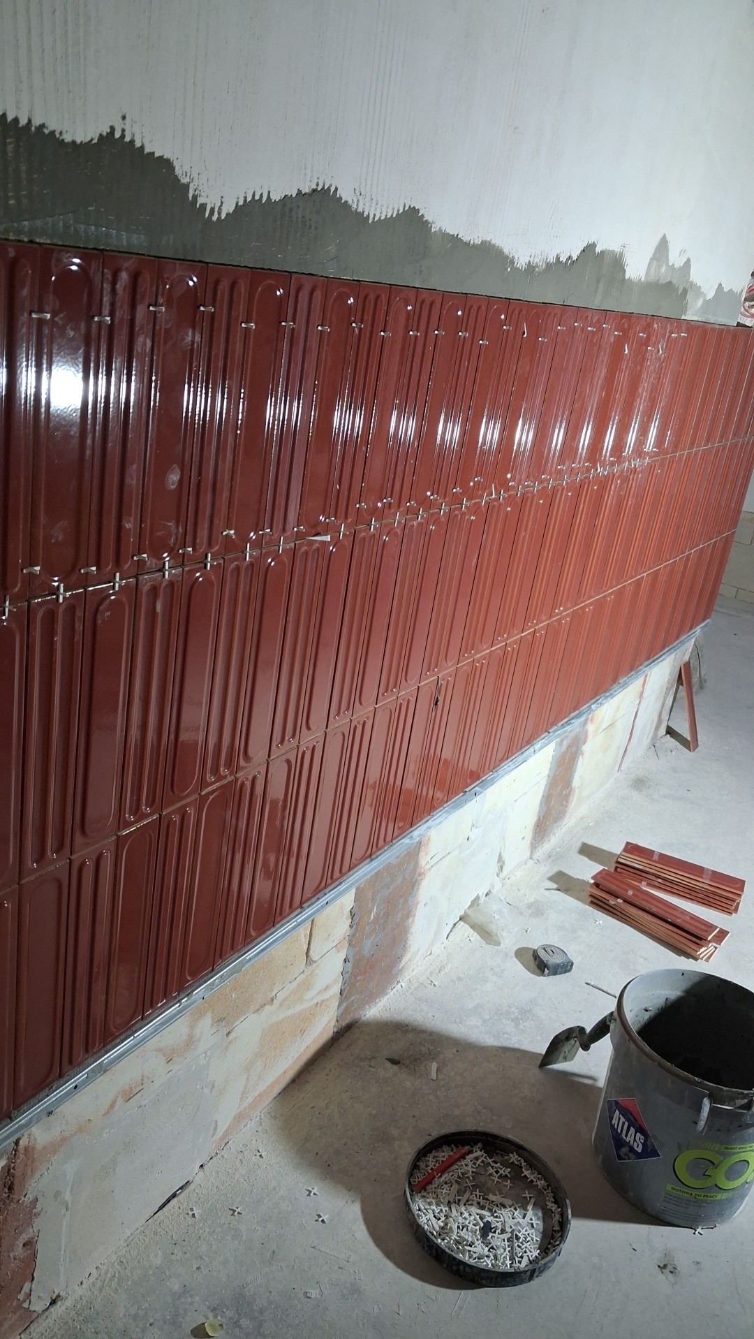 Układanie błyszczących, bordowych płytek na ścianie z widocznym klejem, narzędziami i wiadrem Atlas. Praca w toku, detale montażu płytek.