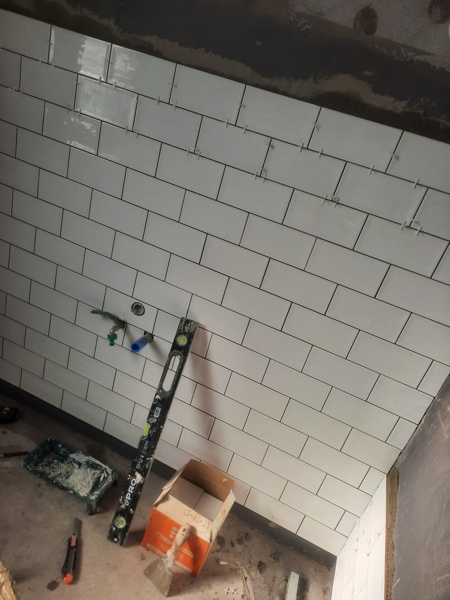 Białe, prostokątne płytki ułożone w cegiełkę na ścianie, z widocznymi krzyżykami dystansowymi, poziomica i narzędzia glazurnicze w tle. Ujęcie z dołu.