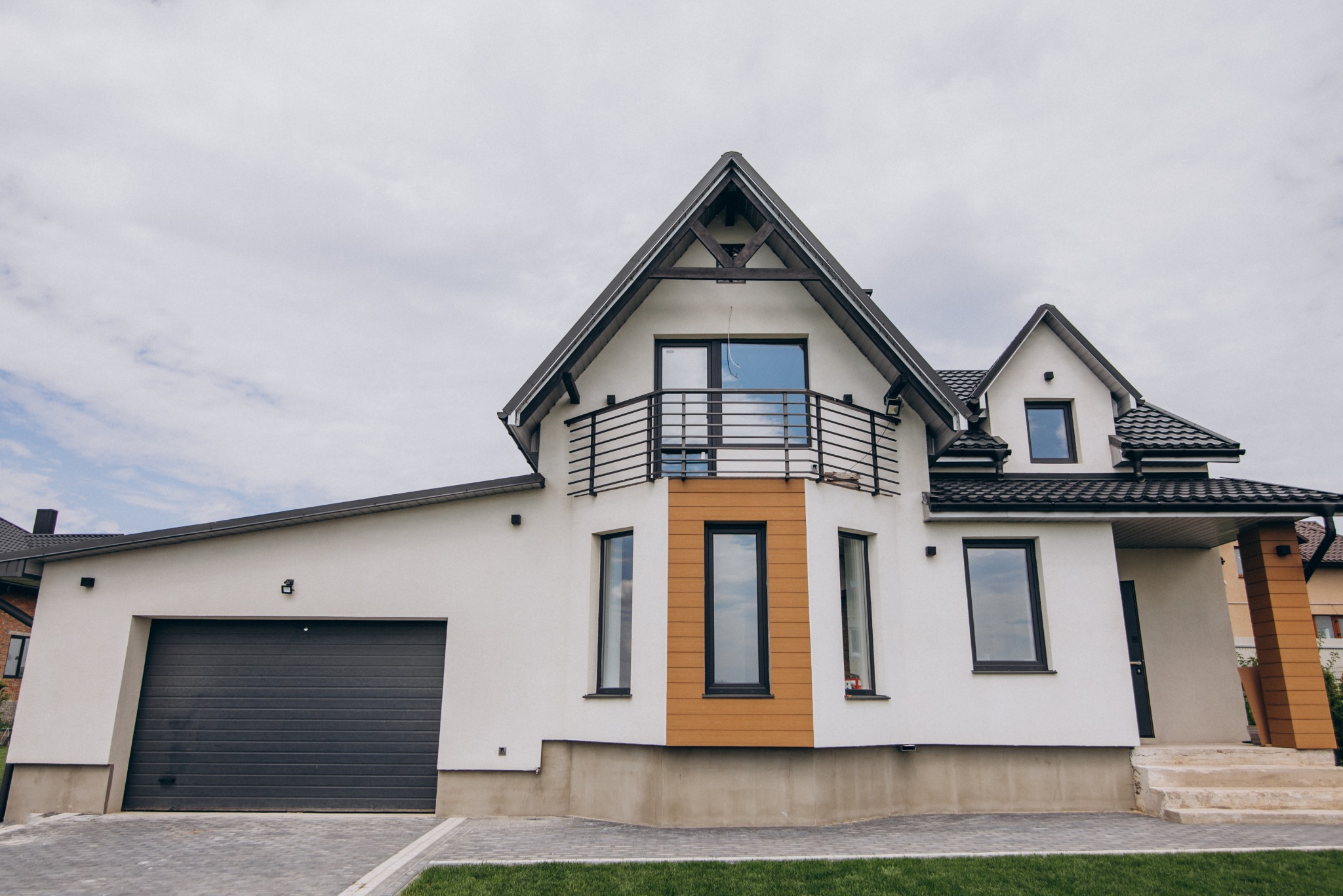 Nowoczesny dom z białymi ścianami, ciemną stolarką okienną i garażem. Uwagę zwraca balkon z czarną balustradą i element dekoracyjny z drewnianych paneli.