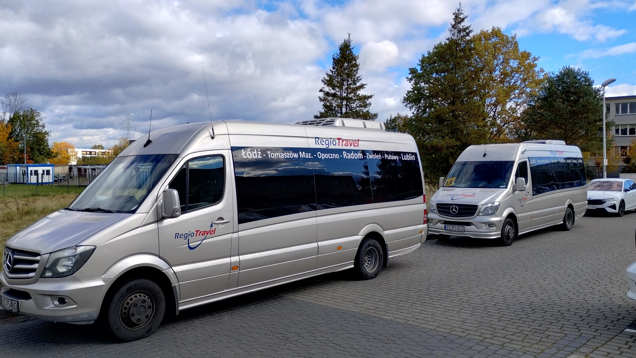 Dwa srebrne busy RegioTravel z napisem trasy Łódź-Lublin, zaparkowane na brukowanym placu, z budynkiem w tle i fragmentem białego samochodu po prawej.