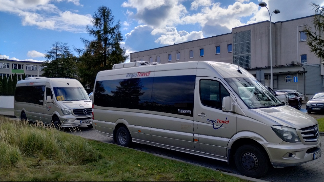 Dwa busy marki Mercedes-Benz Sprinter w kolorze szampańskim z logo RegioTravel zaparkowane przed budynkiem. Jeden z busów ma naklejkę z sylwetkami dzieci.