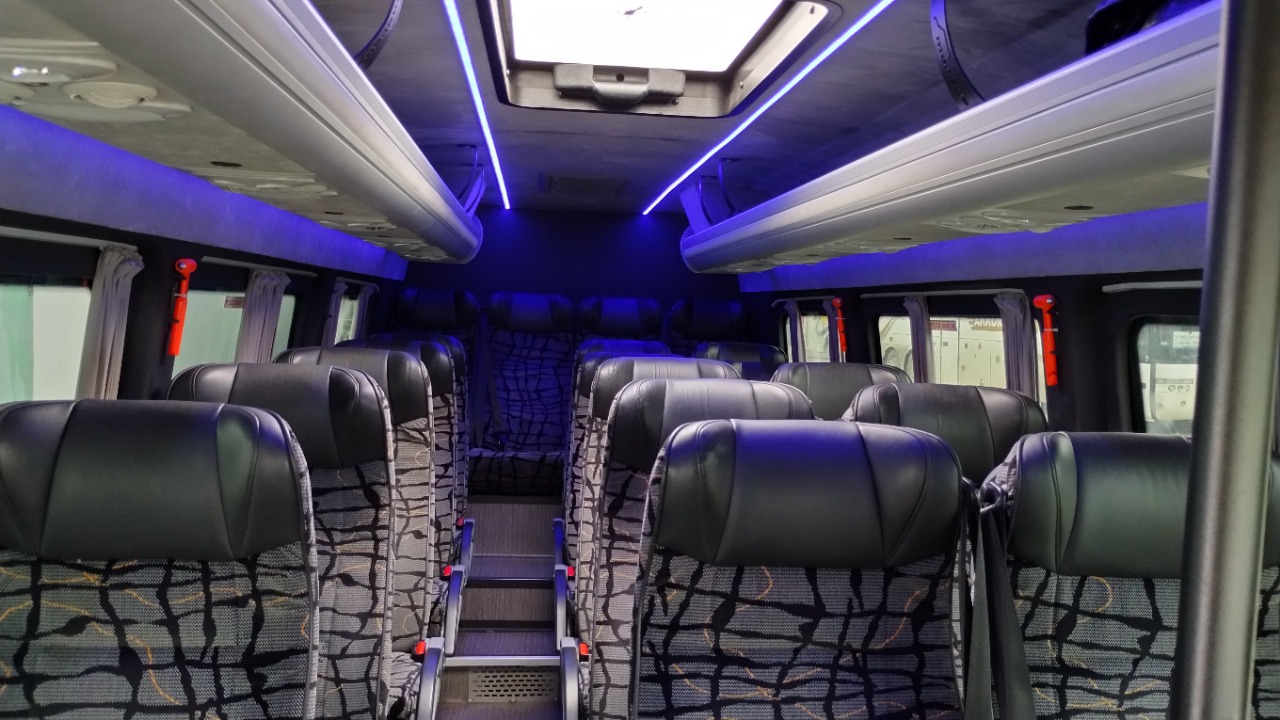 Wnętrze busa z rzędami czarnych foteli z wzorzystą tapicerką, oświetlone niebieskim światłem LED. Widoczne okna z zasłonami i schodki.