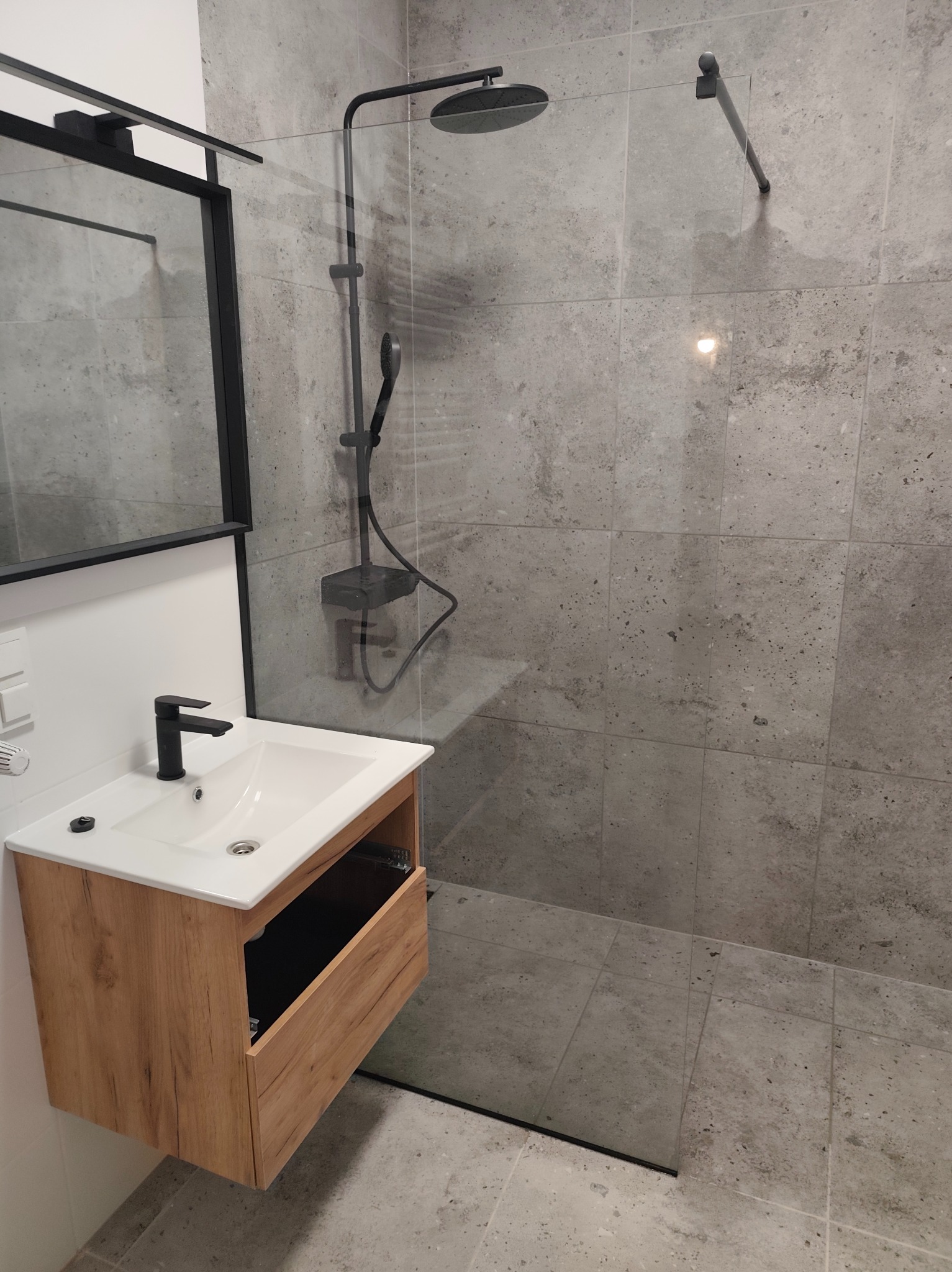 Nowoczesna łazienka z kabiną typu walk-in, szarą glazurą i drewnianą szafką pod umywalką z czarną armaturą. Minimalistyczny design, oświetlenie punktowe.