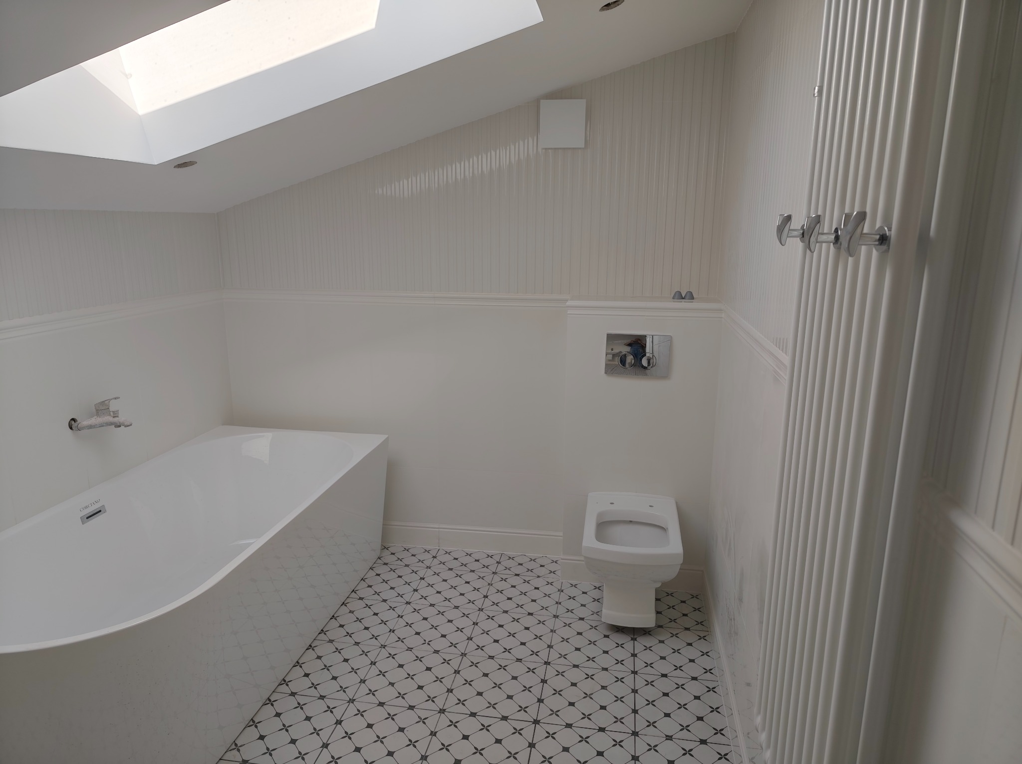 Biała łazienka na poddaszu z wanną, toaletą i grzejnikiem żeberkowym. Uwagę przykuwają wzorzyste płytki podłogowe i okno dachowe wpuszczające naturalne światło.