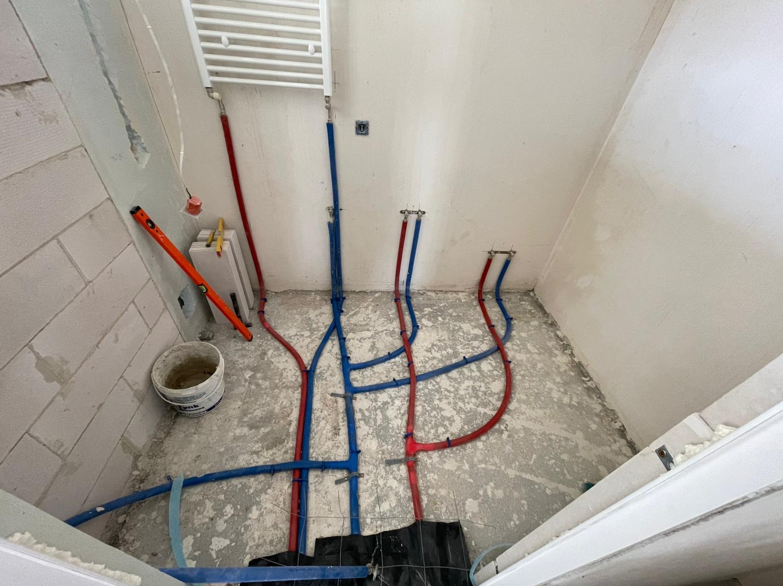 Łazienka w trakcie remontu: widoczne rury instalacji hydraulicznej w kolorze niebieskim i czerwonym, grzejnik drabinkowy, ściany w stanie surowym, poziomica.