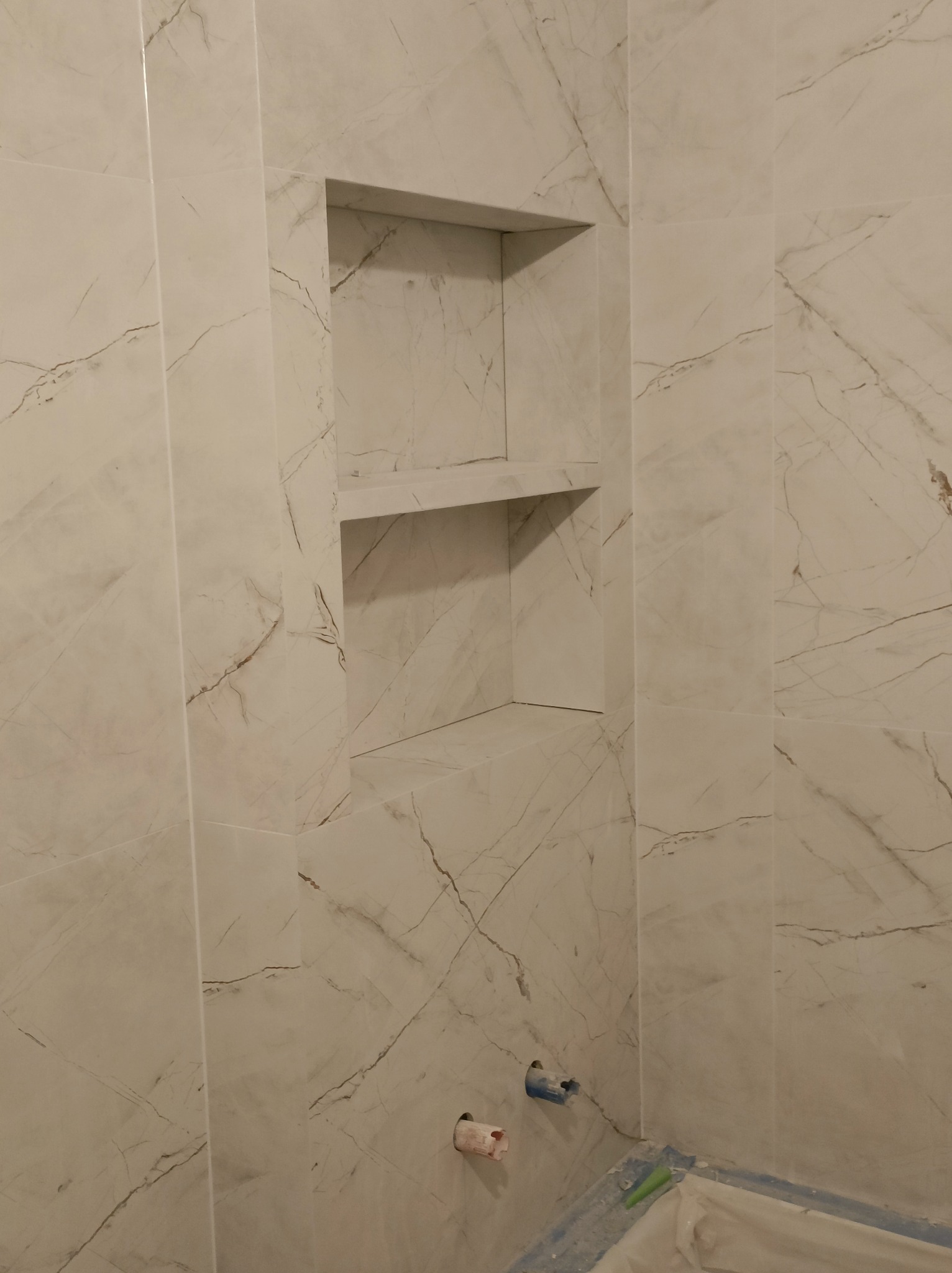 Wykończona ściana łazienki z płytek imitujących marmur, z wnęką na kosmetyki. Widoczne fragmenty instalacji hydraulicznej i zabezpieczenia podłogi.