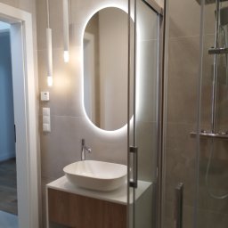 Gabriel Gryglak ProStyle Home Remonty i Wykończenia - Nowoczesna łazienka z owalnym lustrem LED, białą umywalką nablatową na szafce z drewnianym frontem i szklaną kabiną prysznicową. Minimalistyczne oświetlenie.