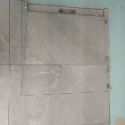 Gabriel Gryglak ProStyle Home Remonty i Wykończenia - Ułożone szare płytki podłogowe z marmurkowym wzorem, widoczna kratka wentylacyjna przy ścianie, niebieska ściana w tle, etap prac wykończeniowych.