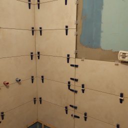Gabriel Gryglak ProStyle Home Remonty i Wykończenia - Ściana w trakcie układania płytek ceramicznych, z użyciem klipsów poziomujących. Widoczne fragmenty instalacji wodnej i przygotowane podłoże.