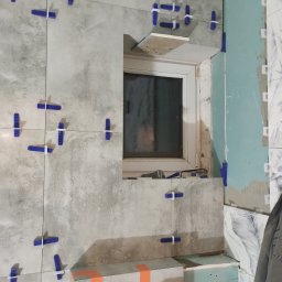 Gabriel Gryglak ProStyle Home Remonty i Wykończenia - Ściana z szarych płytek w trakcie układania, z niebieskimi krzyżykami dystansowymi, widoczne okno i fragment instalacji. Prace glazurnicze w łazience.