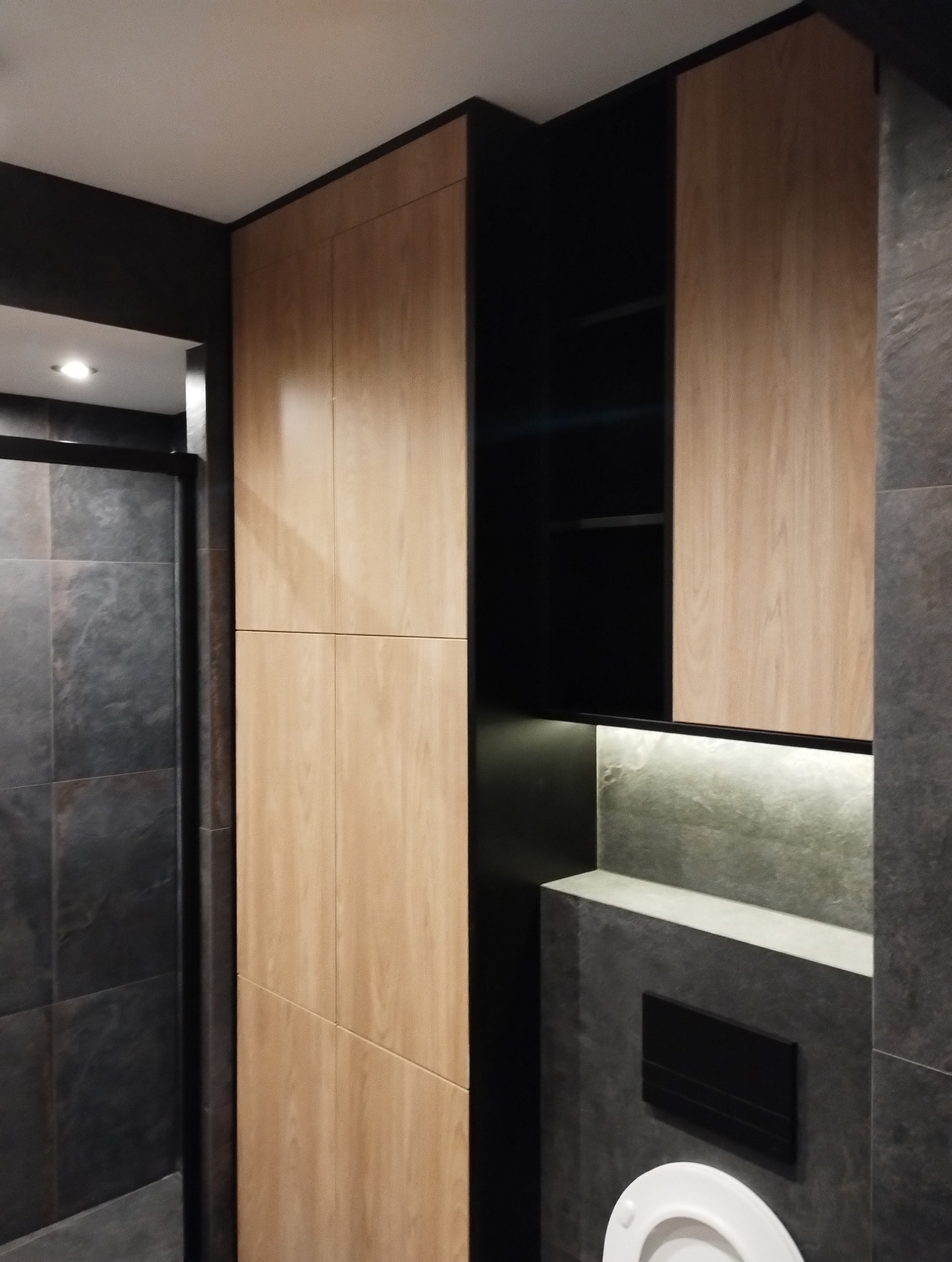 Nowoczesna łazienka z drewnianą zabudową szafek, szarą glazurą i podświetleniem LED nad toaletą. Minimalistyczny design z akcentami drewna i kamienia.
