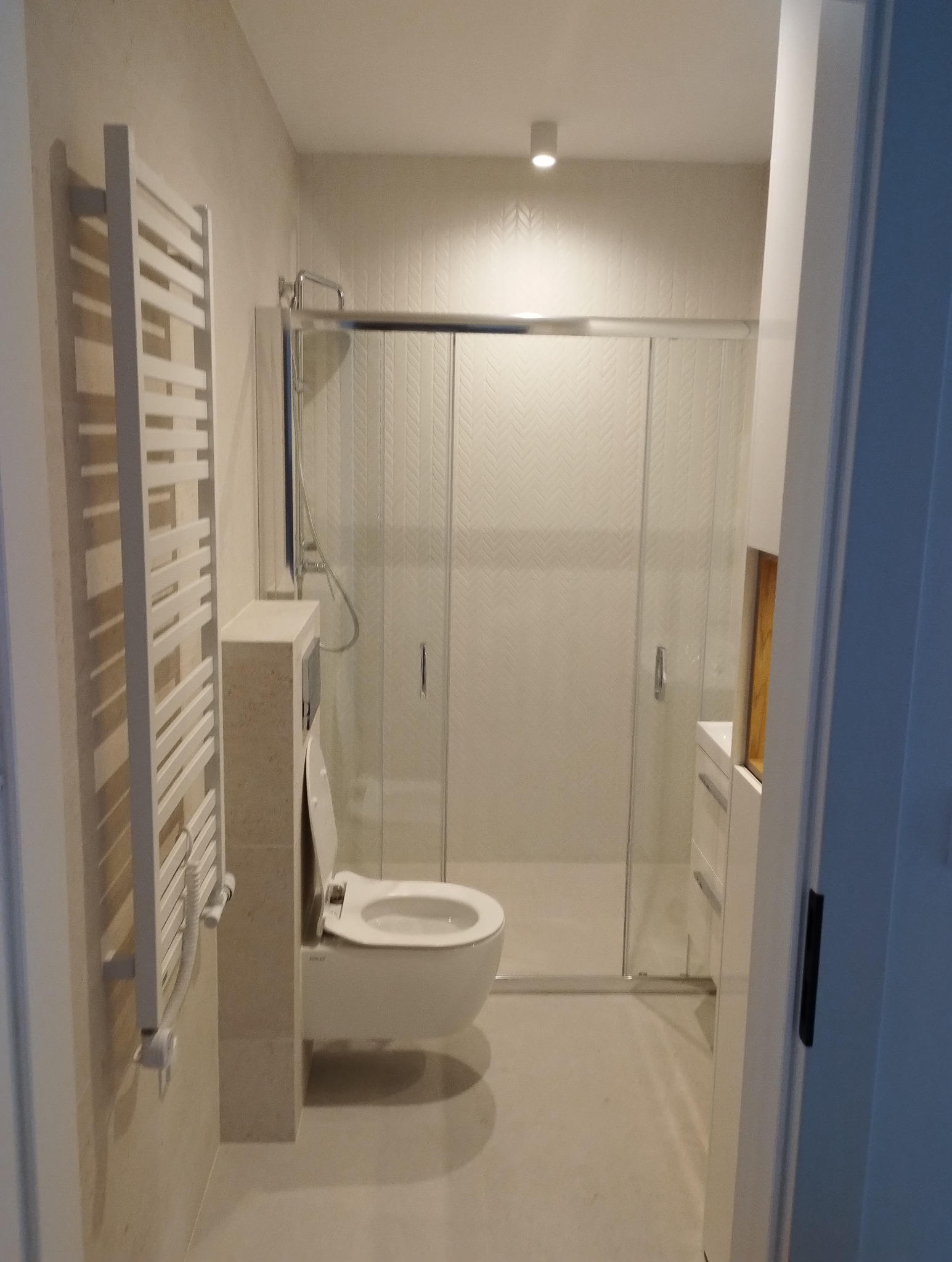 Jasna łazienka po remoncie: kabina prysznicowa ze szklanymi drzwiami, podwieszana toaleta, grzejnik drabinkowy i subtelne wzory na ścianach.