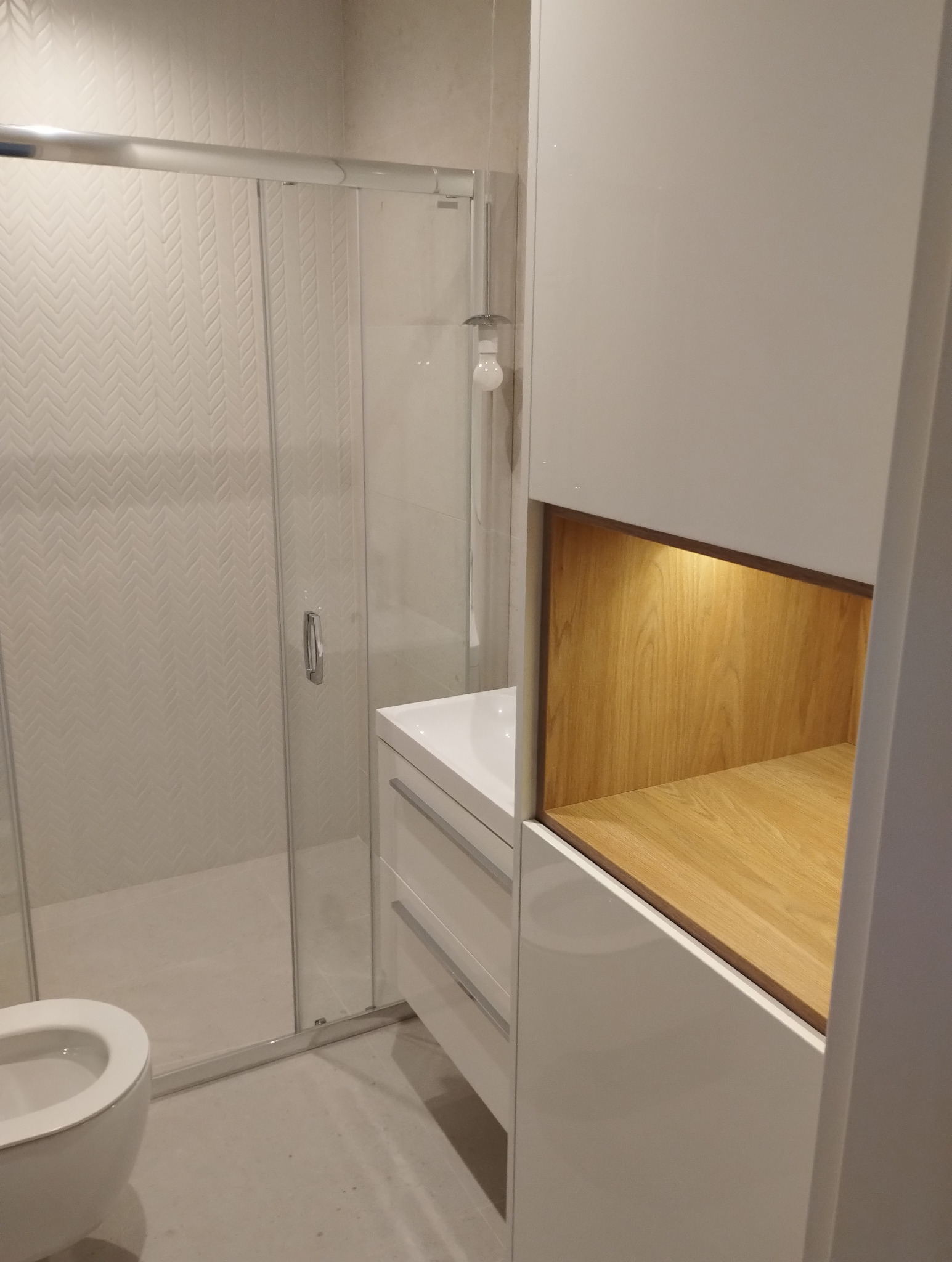 Nowoczesna łazienka z kabiną prysznicową i białą szafką z drewnianą wnęką. Ściany wyłożone płytkami w subtelny wzór jodełki. Minimalistyczny design.