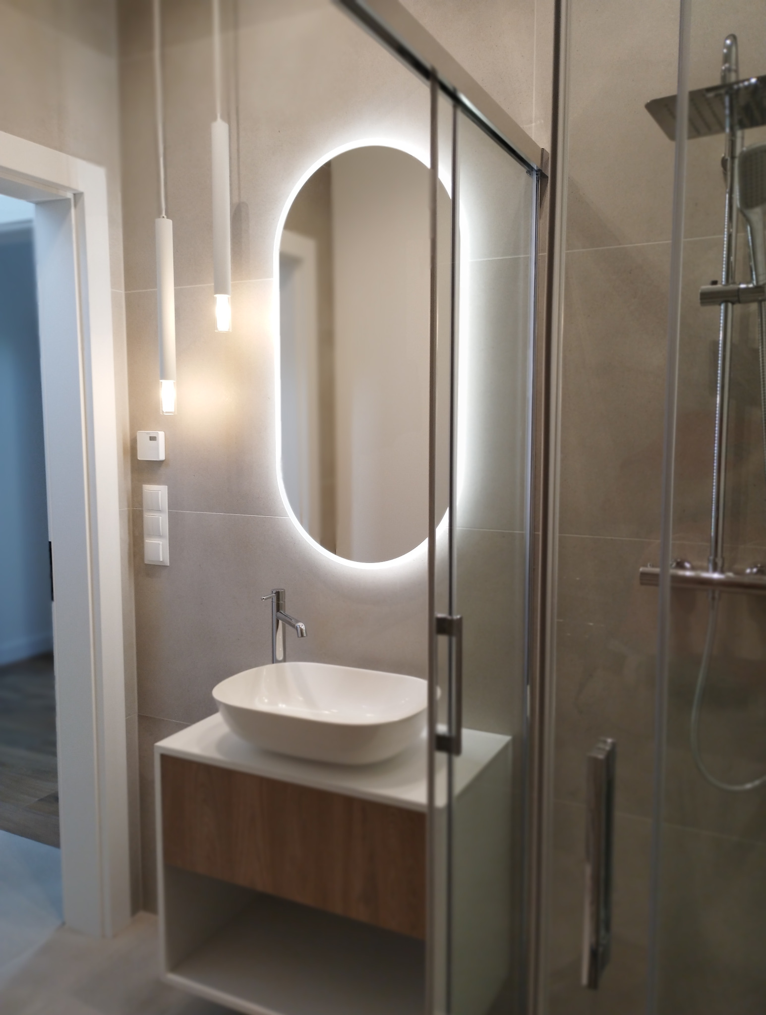 Nowoczesna łazienka z owalnym lustrem LED, białą umywalką nablatową na szafce z drewnianym frontem i szklaną kabiną prysznicową. Minimalistyczne oświetlenie.
