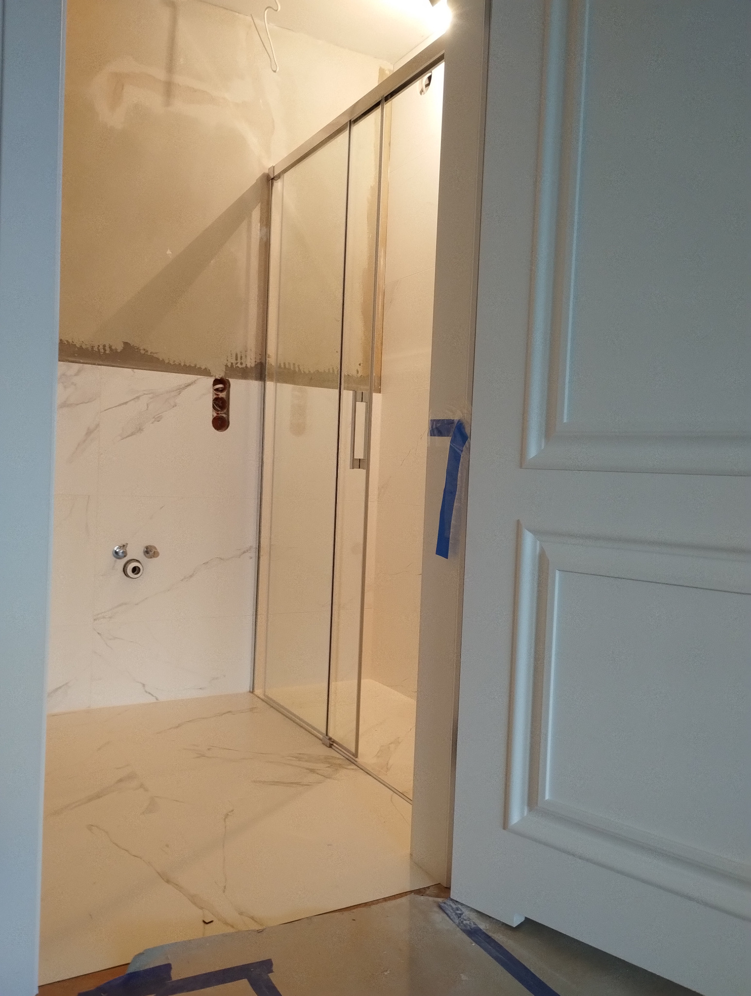 Wnętrze łazienki w trakcie remontu: widoczna kabina prysznicowa ze szklanymi drzwiami, marmurowe płytki na ścianach i podłodze, oraz białe drzwi z niebieską taśmą zabezpieczającą.