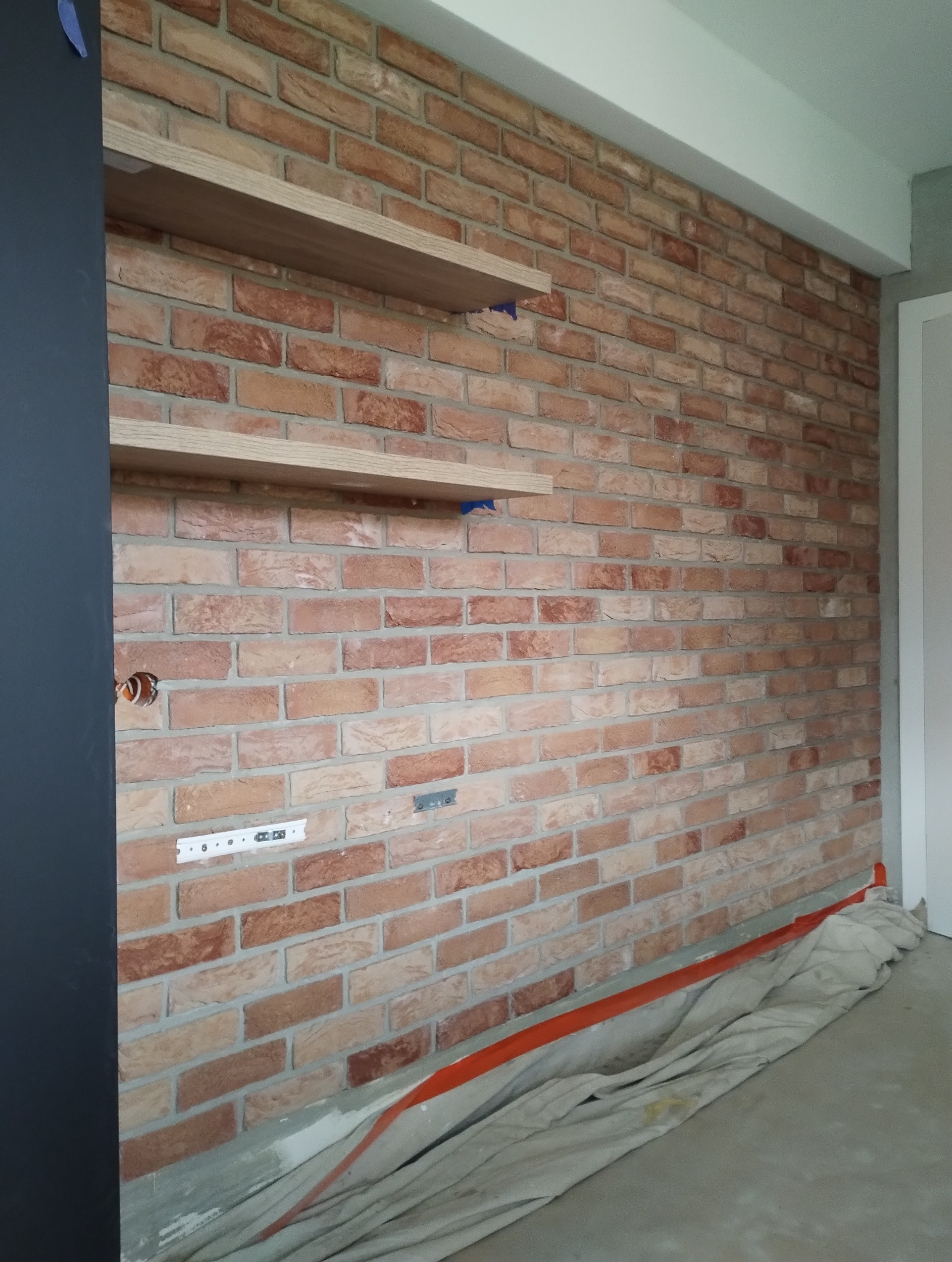 Ściana z cegły klinkierowej z drewnianymi półkami, zabezpieczona folią malarską. Widoczne gniazdko elektryczne i listwa montażowa. Wykończenie wnętrza w stylu industrialnym.