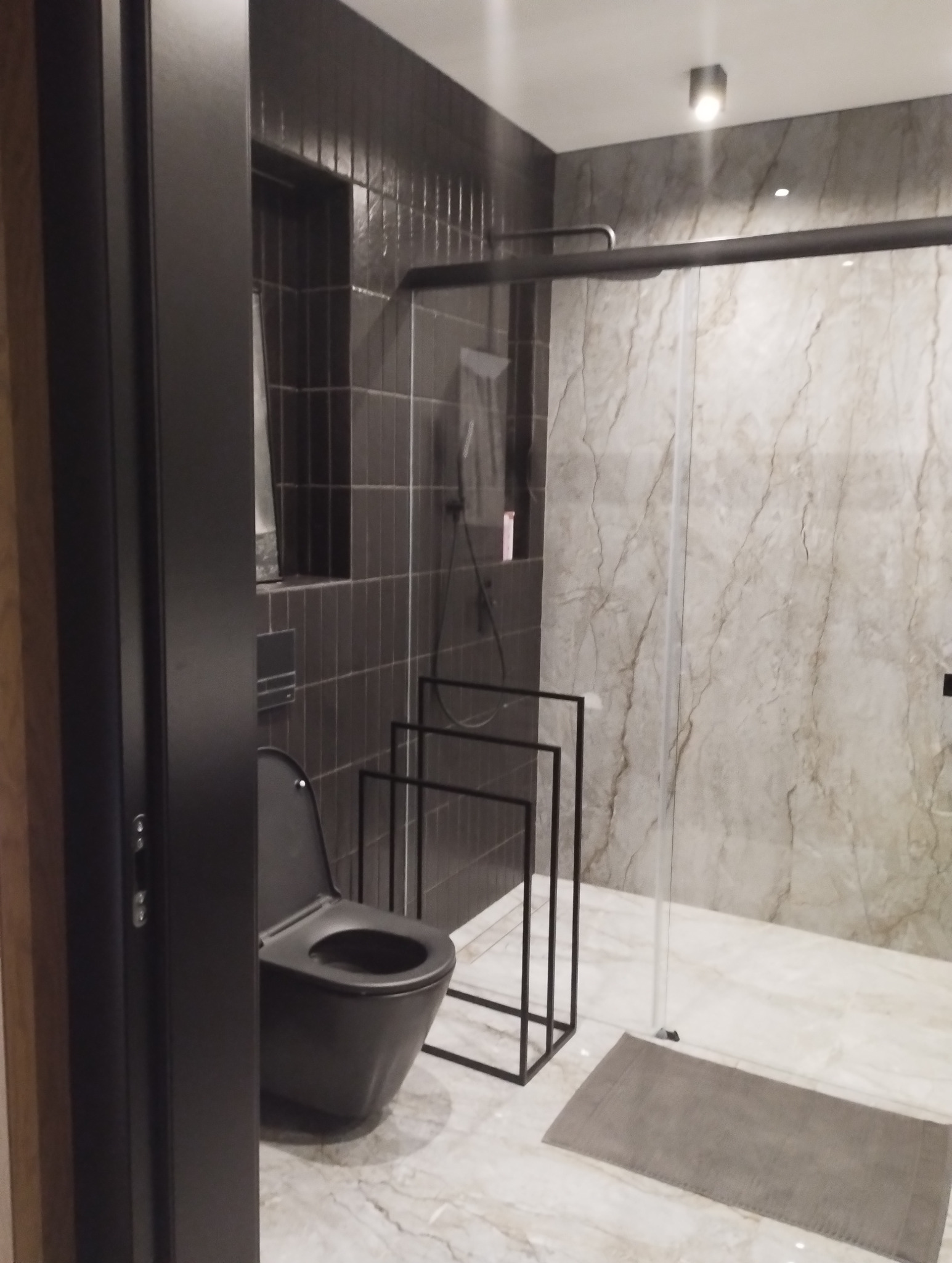 Nowoczesna łazienka z czarną toaletą i kabiną prysznicową z marmurowymi płytkami. Minimalistyczny design z akcentami czerni i bieli. Widoczny fragment drzwi.