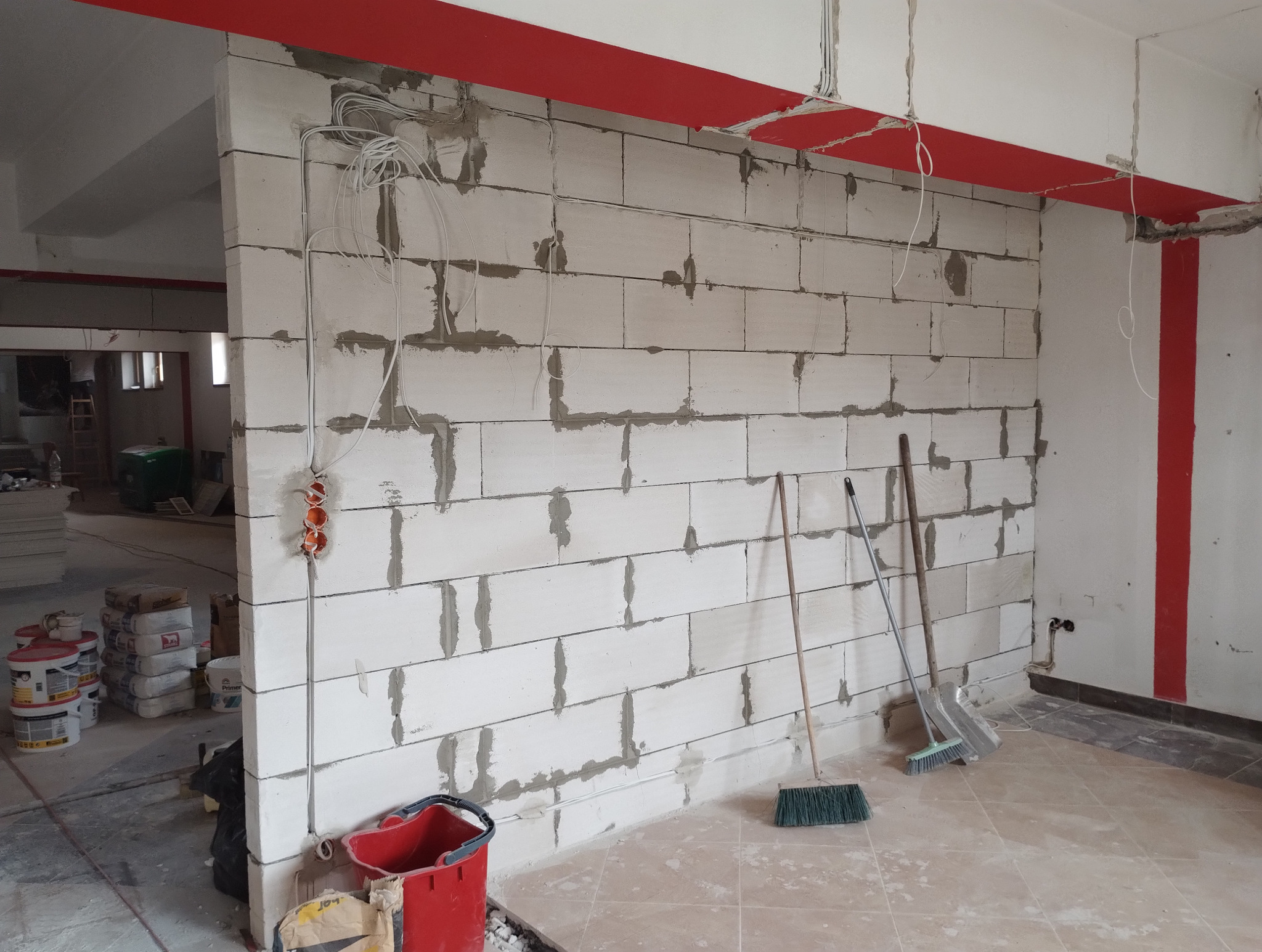 Ściana z białych bloczków z widocznymi spoinami, instalacja elektryczna, narzędzia budowlane i materiały wykończeniowe w surowym wnętrzu z czerwoną belką stropową.
