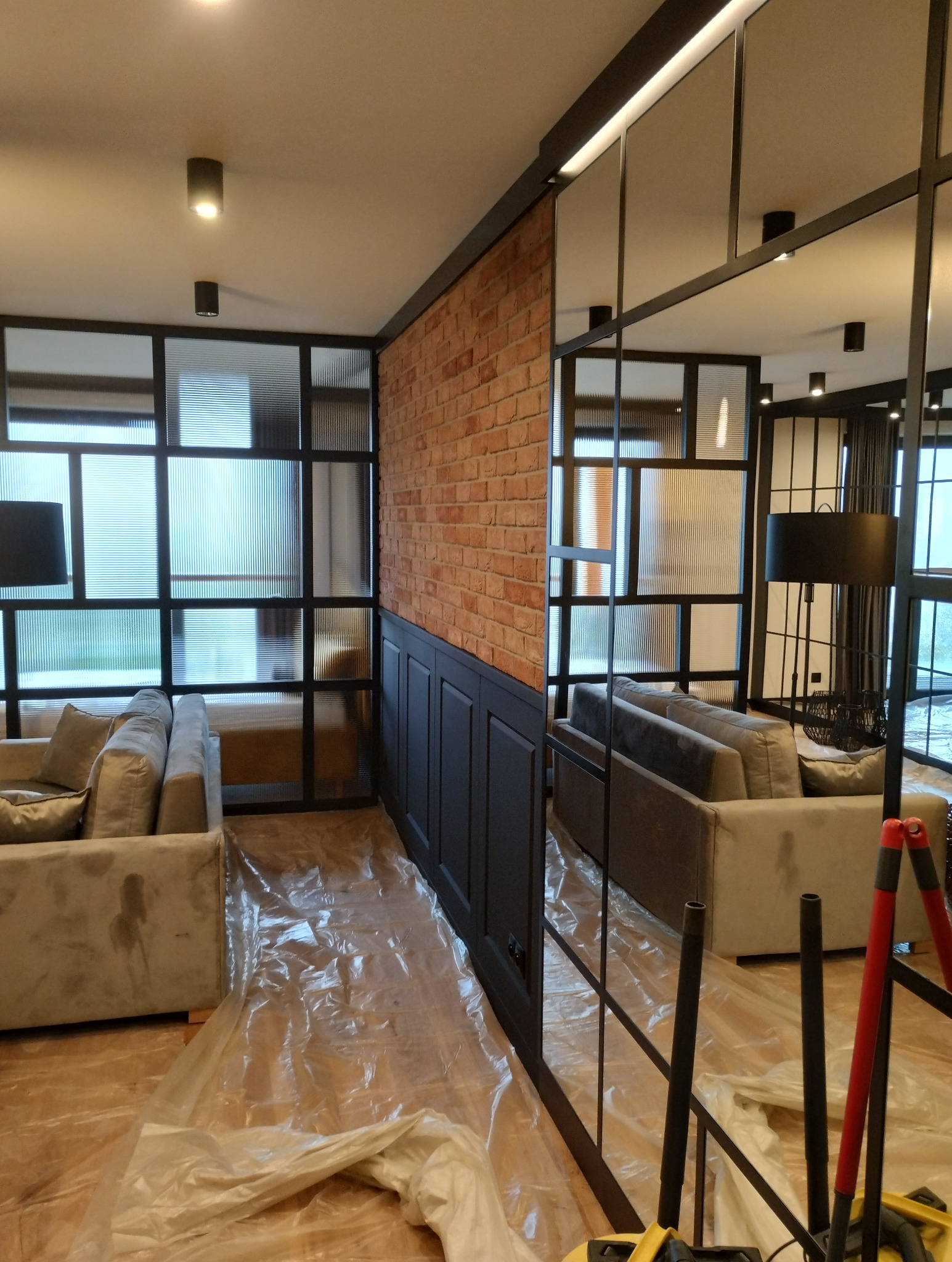 Salon po metamorfozie: ceglana ściana, czarna boazeria, szklana ścianka działowa z czarnymi szprosami. Podłoga zabezpieczona folią malarską.