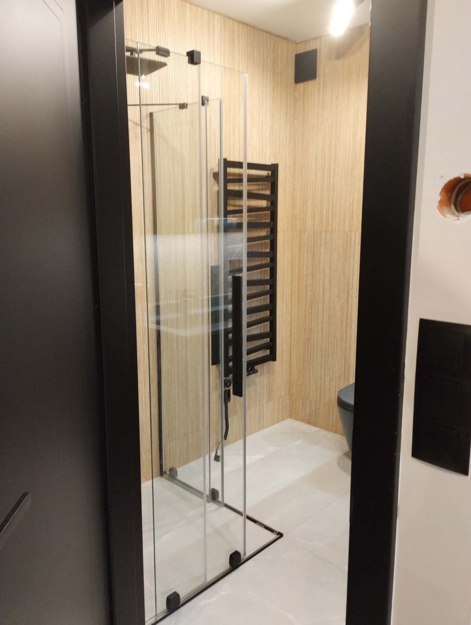 Nowoczesna łazienka po remoncie: kabina prysznicowa ze szkła z czarnymi profilami, grzejnik drabinkowy na ścianie z okładziną imitującą drewno, białe płytki podłogowe.