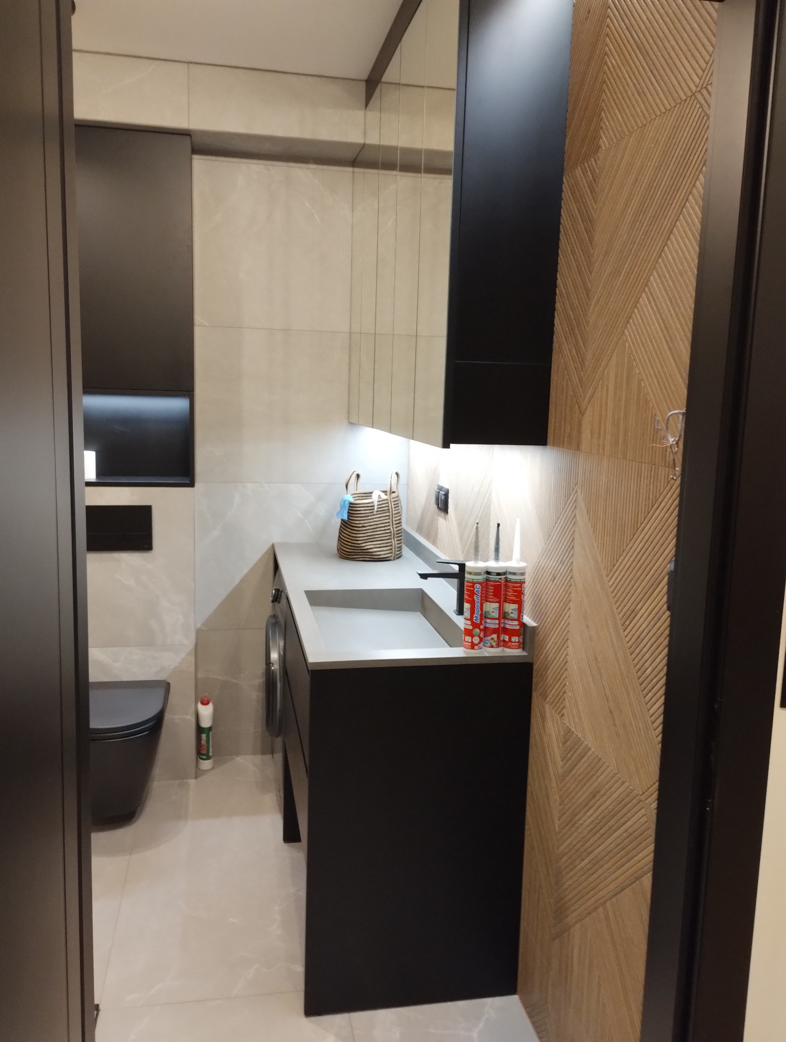 Nowoczesna łazienka w Łapszach Wyżnych z czarną armaturą, szarą umywalką i drewnianą ścianą z geometrycznym wzorem. Widoczne silikony budowlane.